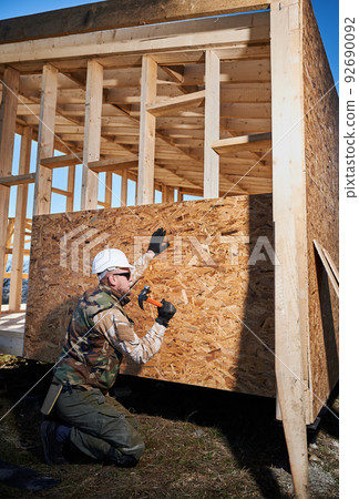 Carpenter hammering nail into OSB panel on the wall of future cottage. Man worker building wooden frame house. Carpentry and construction concept. 92690092