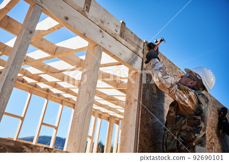 Carpenter hammering nail into OSB panel on the wall of future cottage. Man worker building wooden frame house. Carpentry and construction concept. 92690101