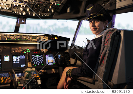 Portrait of woman copilot in uniform flying airplane jet from cockpit with control panel command and dashboard buttons, aerial transportation. Woman using switch and lever with radar navigation. Portrait of woman copilot in uniform flying airplane jet from cockpit with control panel command and dashboard buttons, aerial transportation. Woman using switch and lever with radar navigation. 92691180