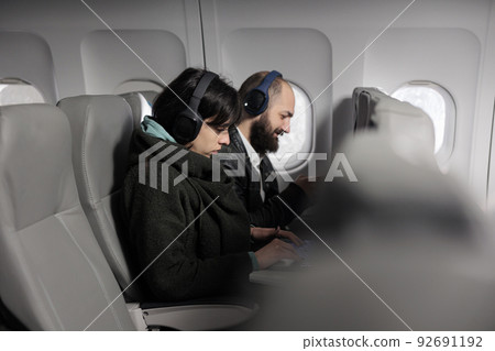 Young travellers using laptop and smartphone on airplane flight, waiting to arrive at holiday vacation. Couple of passengers working on devices and travelling with international airline. 92691192
