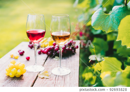 Glasses with white and red wine and grape berries on the wooden table in the vineyards, winery with green leaves background in sunset light. Wine tasting, Degustation. Selective focus, Copy space 92693828