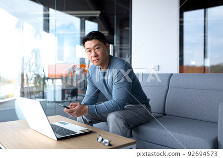Serious and focused freelance businessman, in modern office, asian man kidnaps in camera, working on laptop, holding phone 92694573