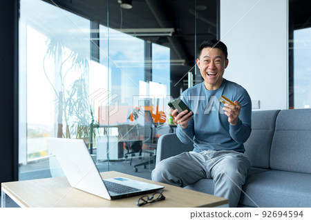 Happy asian male businessman holding credit card and phone, online shopping worker sitting in office 92694594