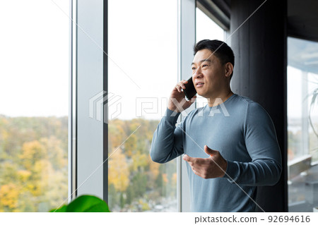Cheerful successful and happy businessman talking in the office on the phone, Asian boss at the big window 92694616