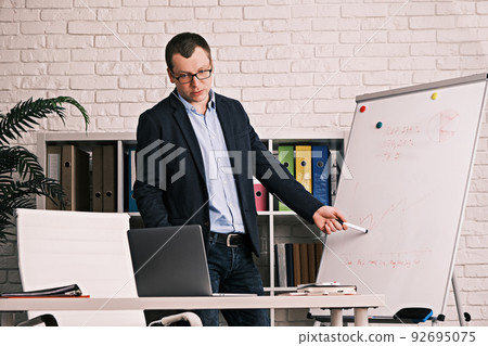 Businessman conducts a presentation online having a video conference. Serious man in glasses and a jacket shows a development graph on a white board while in an office or home office. Businessman conducts a presentation online having a video conference. Serious man in glasses and a jacket shows a development graph on a white board while in an office or home office. 92695075