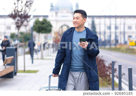 Asian tourist, male passenger arrives from the airport, walks around the city with a suitcase, a man smiles and rejoices uses the phone and the application to book accommodation and book a taxi Asian tourist, male passenger arrives from the airport, walks around the city with a suitcase, a man smiles and rejoices uses the phone and the application to book accommodation and book a taxi 92695185