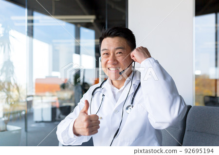 Happy doctor smiles and looks at the camera, Asian announces good news to the patient, uses a video call, and a headset online meeting with colleagues, fun gesturing with his hands, webcam view Happy doctor smiles and looks at the camera, Asian announces good news to the patient, uses a video call, and a headset online meeting with colleagues, fun gesturing with his hands, webcam view 92695194