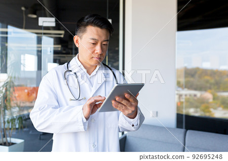 Asian doctor works online with a tablet computer in a modern clinic office, provides advice to online patients, conducts correspondence, discussion, thoughtful men 92695248