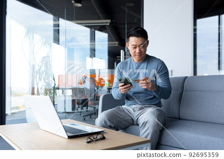 Successful and happy Asian makes online purchases with a credit card and an application on the phone, a businessman spends time in a modern office on a laptop 92695359