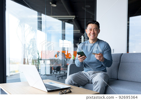 Successful and happy Asian makes online purchases with a credit card and an application on the phone, a businessman spends time in a modern office on a laptop Successful and happy Asian makes online purchases with a credit card and an application on the phone, a businessman spends time in a modern office on a laptop 92695364