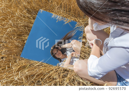 Young woman is iooking at her reflection in the mirrorin a wheat field Young woman is iooking at her reflection in the mirrorin a wheat field 92695570
