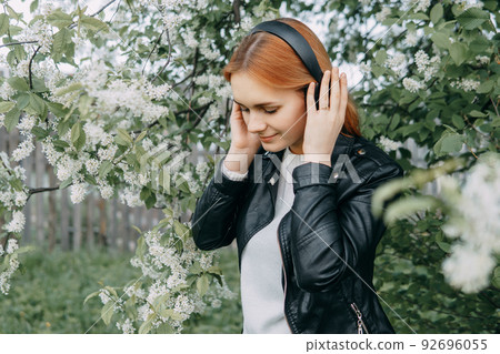 A red-haired teenage girl listens to music in headphones, configured via the phone. The time of spring flowering of cherry orchards A red-haired teenage girl listens to music in headphones, configured via the phone. The time of spring flowering of cherry orchards 92696055