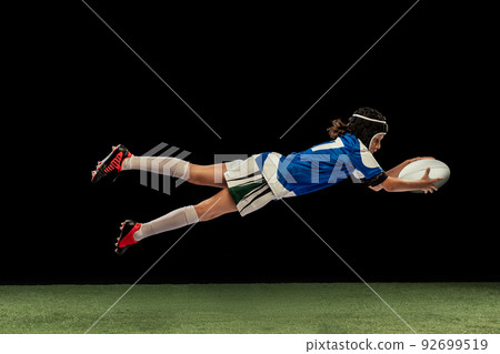Dynamic portrait of school age boy, junior male rugby player practicing rugby football isolated on dark background with grass floooring. Dynamic portrait of school age boy, junior male rugby player practicing rugby football isolated on dark background with grass floooring. 92699519