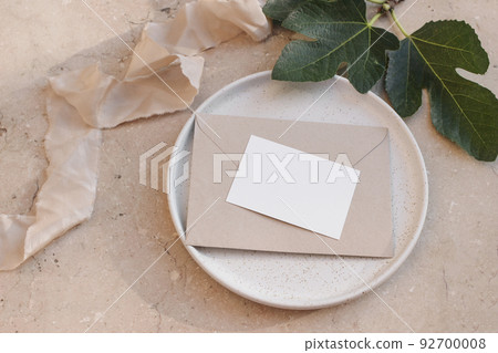 Summer branding, wedding stationery. Mediterranean design. Business card, invitation mock up on craft envelope and ceramic plate. Green fig leaves and silk ribbon. Beige marble background. Flatlay Summer branding, wedding stationery. Mediterranean design. Business card, invitation mock up on craft envelope and ceramic plate. Green fig leaves and silk ribbon. Beige marble background. Flatlay 92700008