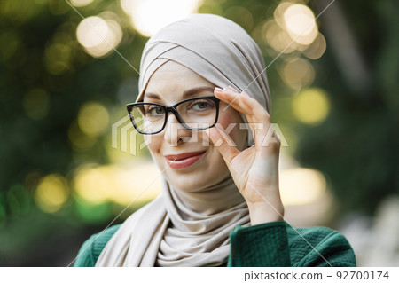 Pretty smiling muslim woman in hijab walks at green summer city park and touching her eyeglass. 92700174