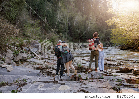 Rear on two couples standing and hugging at river on rocks. Hikers in mountains valley. 92706543