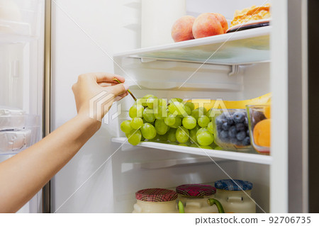 Woman hand taking, grabbing or picks up green bunch of grapes out of open refrigerator shelf or fridge drawer full of fruits, blueberries, bottles with yogurt. Healthy food diet, lifestyle concept Woman hand taking, grabbing or picks up green bunch of grapes out of open refrigerator shelf or fridge drawer full of fruits, blueberries, bottles with yogurt. Healthy food diet, lifestyle concept 92706735