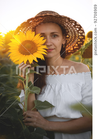 A young girl in a white dress and hat in a field of sunflowers at sunset. Portrait of a woman with a slim figure on a background of yellow flowers 92708989