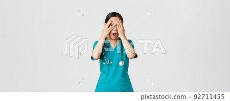 Covid-19, healthcare workers and preventing virus concept. Scared and shocked asian female nurse, doctor in scrubs witness something embarrassing or scary, cover eyes but peek through fingers 92711455