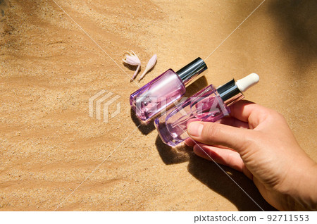 Close-up of the hand putting skin care beauty product packaging in a light violet glass bottle on golden sand background Close-up of the hand putting skin care beauty product packaging in a light violet glass bottle on golden sand background 92711553