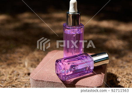 Light purple dropper bottles on pink stone, on fallen dry pine trees needles, with sunlight and coniferous trees shadow 92711563
