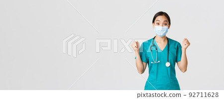 Covid-19, coronavirus disease, healthcare workers concept. Winning cheerful asian female doctor, physician in medical mask and scrubs, look upbeat, rejoicing, say hooray, white background 92711628