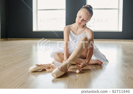 Youthful blond ballerina in white tutu and beige leggins tying her pointe shoes Youthful blond ballerina in white tutu and beige leggins tying her pointe shoes 92711840
