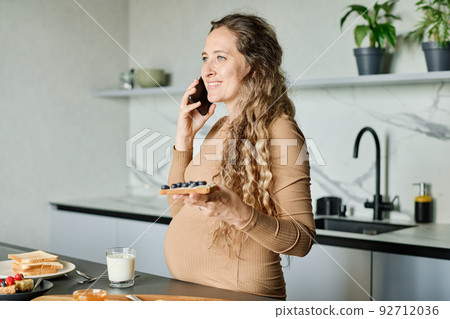Young cheerful pregnant woman with sandwich communicating on smartphone 92712036
