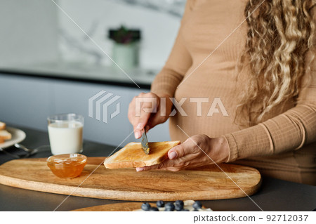 Hands of young pregnant woman with knife spreading honey or jam on bread 92712037