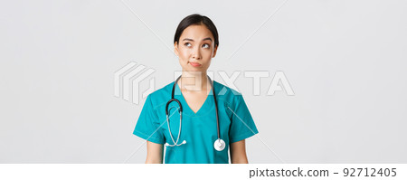 Healthcare workers, preventing virus, quarantine campaign concept. Thoughtful asian female doctor, nurse in scrubs looking upper left corner and smirk unsure, thinking, white background Healthcare workers, preventing virus, quarantine campaign concept. Thoughtful asian female doctor, nurse in scrubs looking upper left corner and smirk unsure, thinking, white background 92712405