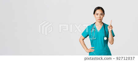 Covid-19, healthcare workers, pandemic concept. Serious-looking professional female asian doctor in scrubs, physician shaking finger in prohibition, warning gesture, forbid something Covid-19, healthcare workers, pandemic concept. Serious-looking professional female asian doctor in scrubs, physician shaking finger in prohibition, warning gesture, forbid something 92712837