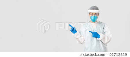 Covid-19, coronavirus disease, healthcare workers concept. Surprised female asian doctor, physician in medical respirator and personal protective equipment pointing upper left corner 92712839