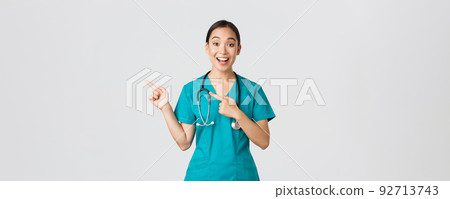 Covid-19, healthcare workers, pandemic concept. Excited asian nurse, doctor in scrubs and stethoscope, pointing fingers upper left corner. Physician smiling amazed as showing awesome announcement 92713743