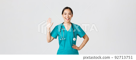 Covid-19, healthcare workers and preventing virus concept. Friendly-looking smiling asian female doctor, physician in scrubs waving hand to say hi, hello, greeting patient, nice to see you 92713751