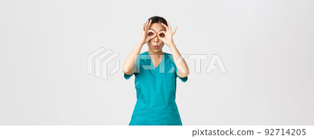 Covid-19, healthcare workers and preventing virus concept. Impressed funny and cute female asian nurse in scrubs looking through fingers over eyes as if stare in binoculats amazed Covid-19, healthcare workers and preventing virus concept. Impressed funny and cute female asian nurse in scrubs looking through fingers over eyes as if stare in binoculats amazed 92714205