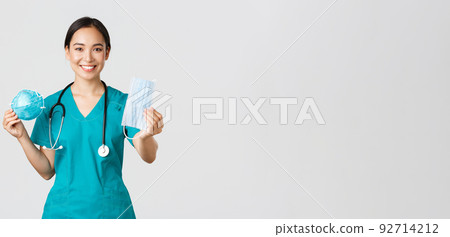 Covid-19, coronavirus disease, healthcare workers concept. Pleasant female physician, asian doctor in scrubs showing respirator and medical mask, provide staff with personal protective equipment 92714212