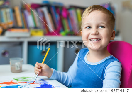 Portrait of cute little blond kid boy enjoy have fun painting with watercolor and brush bright abstract inage on paper at home or kindergarten class. Imagination development children play activity 92714347