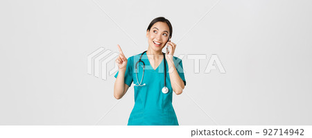 Covid-19, healthcare workers and preventing virus concept. Portrait of young pretty asian female doctor, nurse in scrubs making online order, talking on phone and pointing upper left corner, smiling Covid-19, healthcare workers and preventing virus concept. Portrait of young pretty asian female doctor, nurse in scrubs making online order, talking on phone and pointing upper left corner, smiling 92714942