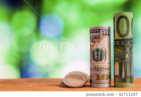 One hundred euro and one hundred US dollar rolled bills banknotes, with euro coins and american cents on green blurred bokeh background. 92715207