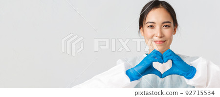 Covid-19, coronavirus disease, healthcare workers concept. Close-up of exhausted happy asian female doctor take-off personal protective equipment, have skin marks from respirator, show heart gesture Covid-19, coronavirus disease, healthcare workers concept. Close-up of exhausted happy asian female doctor take-off personal protective equipment, have skin marks from respirator, show heart gesture 92715534