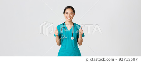 Covid-19, healthcare workers, pandemic concept. Professional confident smiling doctor, female physician in scrubs showing thumbs-up with assured expression, ensure all good, perfect or excellent 92715597