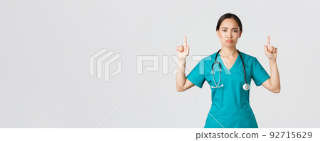 Covid-19, healthcare workers, pandemic concept. Gloomy disappointed asian female doctor, physician in scrubs pointing fingers up and grimacing displeased, standing white background 92715629