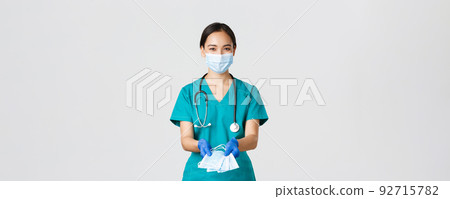 Covid-19, coronavirus disease, healthcare workers concept. Friendly smiling asian physician, female doctor in scrubs and rubber gloves handing over medical masks to patients, white background 92715782