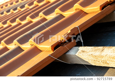 Closeup of metal montage anchor for installation of yellow ceramic roofing tiles mounted on wooden boards covering residential building roof under construction. 92715789