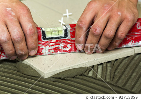 Ceramic tiles and tools for tiler. Worker hand installing floor tiles. Home improvement, renovation - ceramic tile floor adhesive, mortar, level. Ceramic tiles and tools for tiler. Worker hand installing floor tiles. Home improvement, renovation - ceramic tile floor adhesive, mortar, level. 92715859
