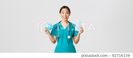Covid-19, coronavirus disease, healthcare workers concept. Amused smiling asian female nurse, inters in scrubs showing respirator and medical mask, receive personal protective equipment Covid-19, coronavirus disease, healthcare workers concept. Amused smiling asian female nurse, inters in scrubs showing respirator and medical mask, receive personal protective equipment 92716159