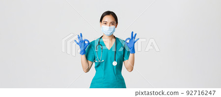 Covid-19, coronavirus disease, healthcare workers concept. Smiling professional and confident asian doctor, physician in medical mask, gloves and scrubs show okay gesture, guarantee quality service 92716247