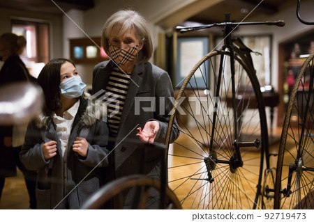 Elderly woman with preteen granddaughter in protective masks examining retro bicycles in museum 92719473
