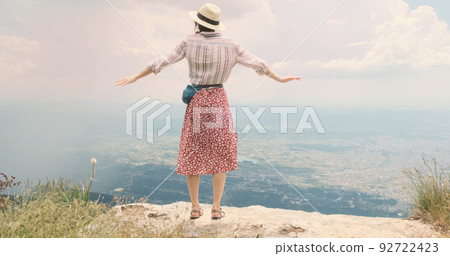 Woman traveler flaps arms like wings on mountain top. Young woman in hat opens arms wide waving like bird's wings over Tira city, Albania. Red dress happy girl blowing wind. Dream, travel concept 92722423