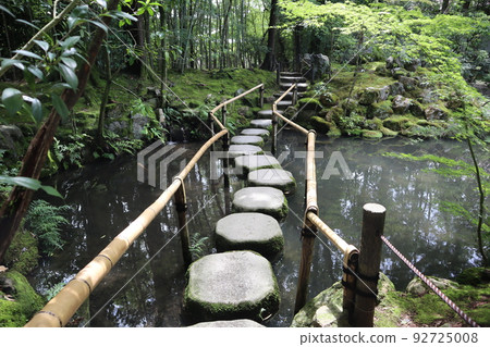 Landscape of a garden surrounded by fresh green trees, green moss and plants with a stepping stone bridge and a pond with bamboo railings 92725008